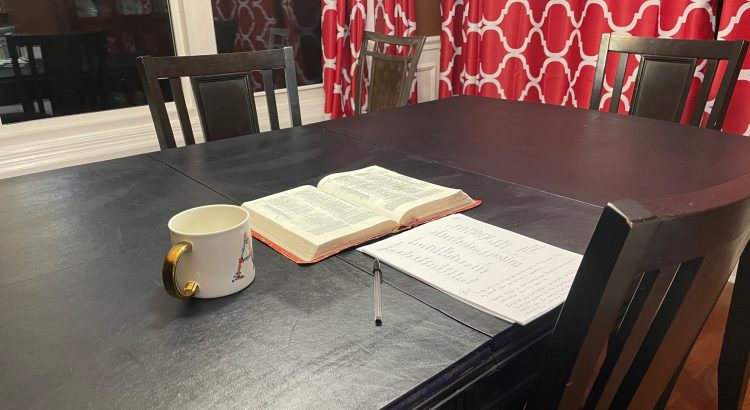 Open Bible, coffee mug, stack of paper on a dining room table for quiet time
